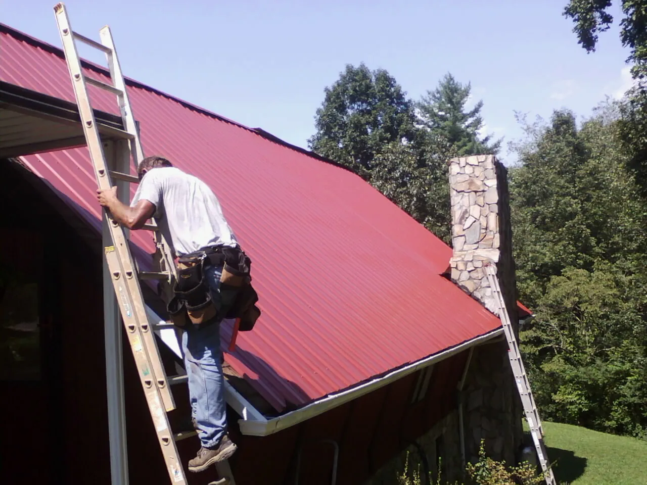 Roofer on a ladder beside a red metal roof and stone chimney — Soffit & Fascia Repair by Roofing Brothers in Milldale